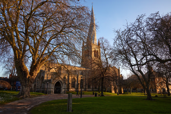 The image shows an architectural photograph of the Church of St Mary and All Saints in Chesterfield, taken on a winter morning. The church is notable for its historic spire and is surrounded by leafless trees, indicating the season. The early morning sunlight casts a golden hue onto the stone façade, highlighting the church's details and the surrounding grounds. This view captures the tranquility of the Chesterfield landmark, with its prominent spire rising above the landscape in the quietness of a winter morning.
